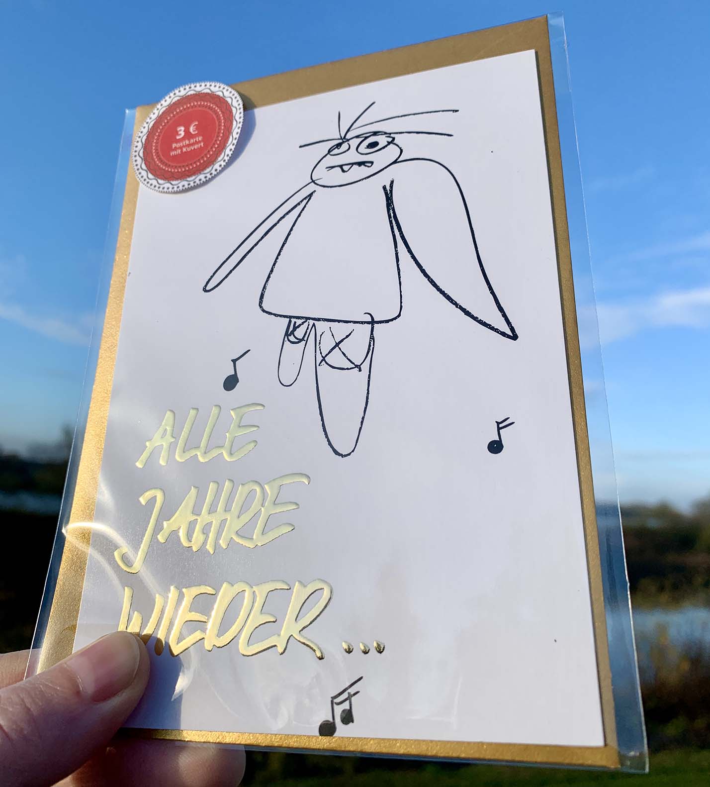 Weihnachtskarte „ ALLE JAHRE WIEDER… müder Engel“ mit goldenem Kuvert – stilvolle und humorvolle Postkarte für Weihnachten und Engel-Liebhaber. Frontansicht mit Klarsichthülle und Sonnenlicht